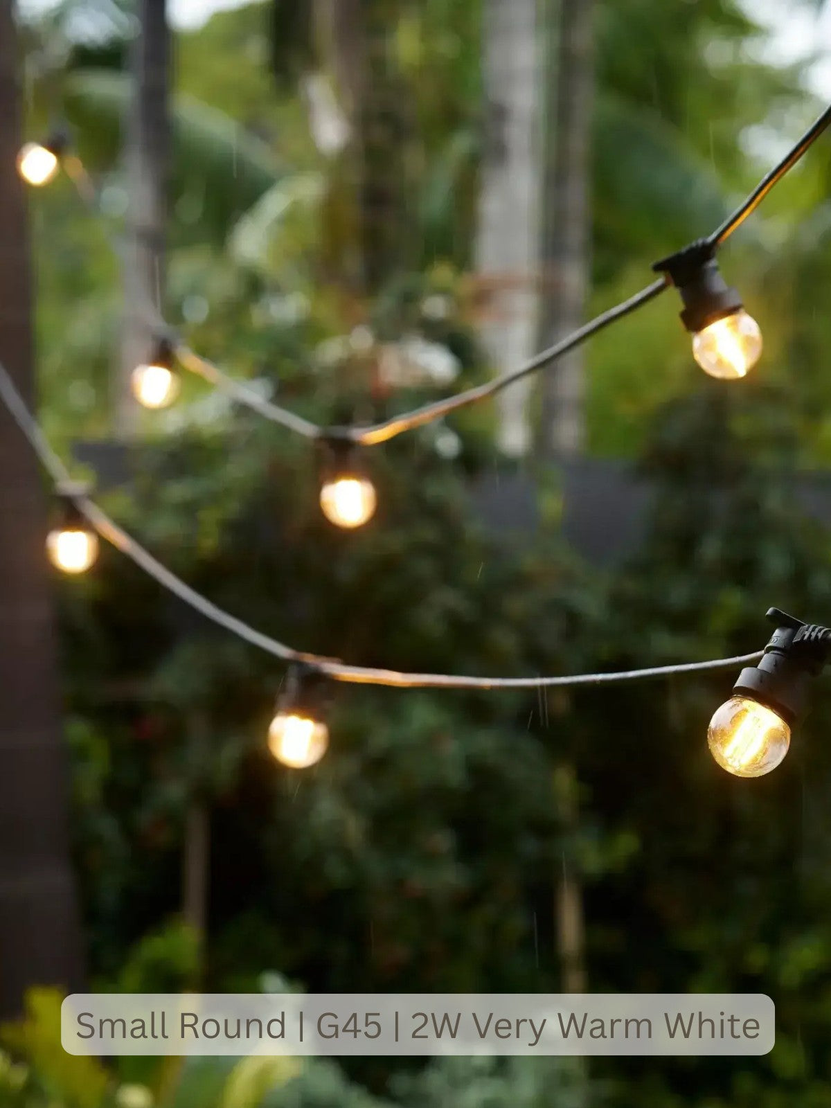 Outdoor string lights with small round warm white bulbs hanging in curved lines against a blurred green garden background. A label at the bottom reads 'Small Round | G45 | 2W Very Warm White'.
