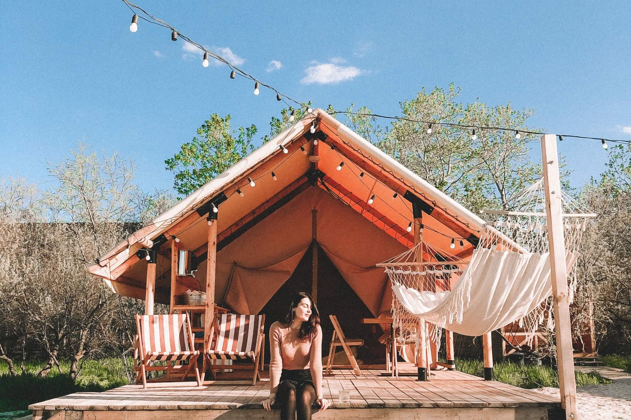 Person sitting on a wooden platform in front of a large tent with striped chairs and a hammock.